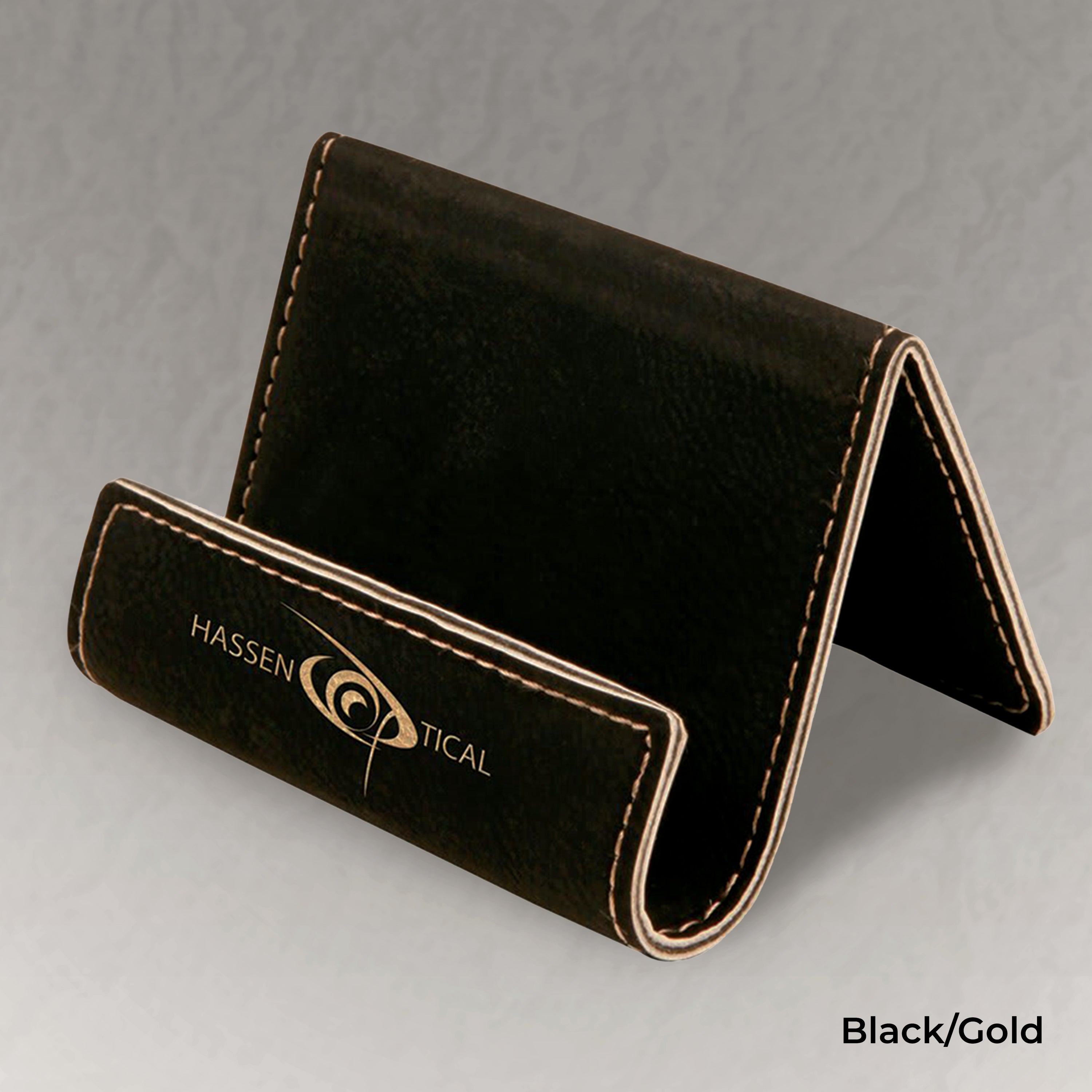Black business card holder with gold stitching on a gray background, featuring the Hassen Tical logo.