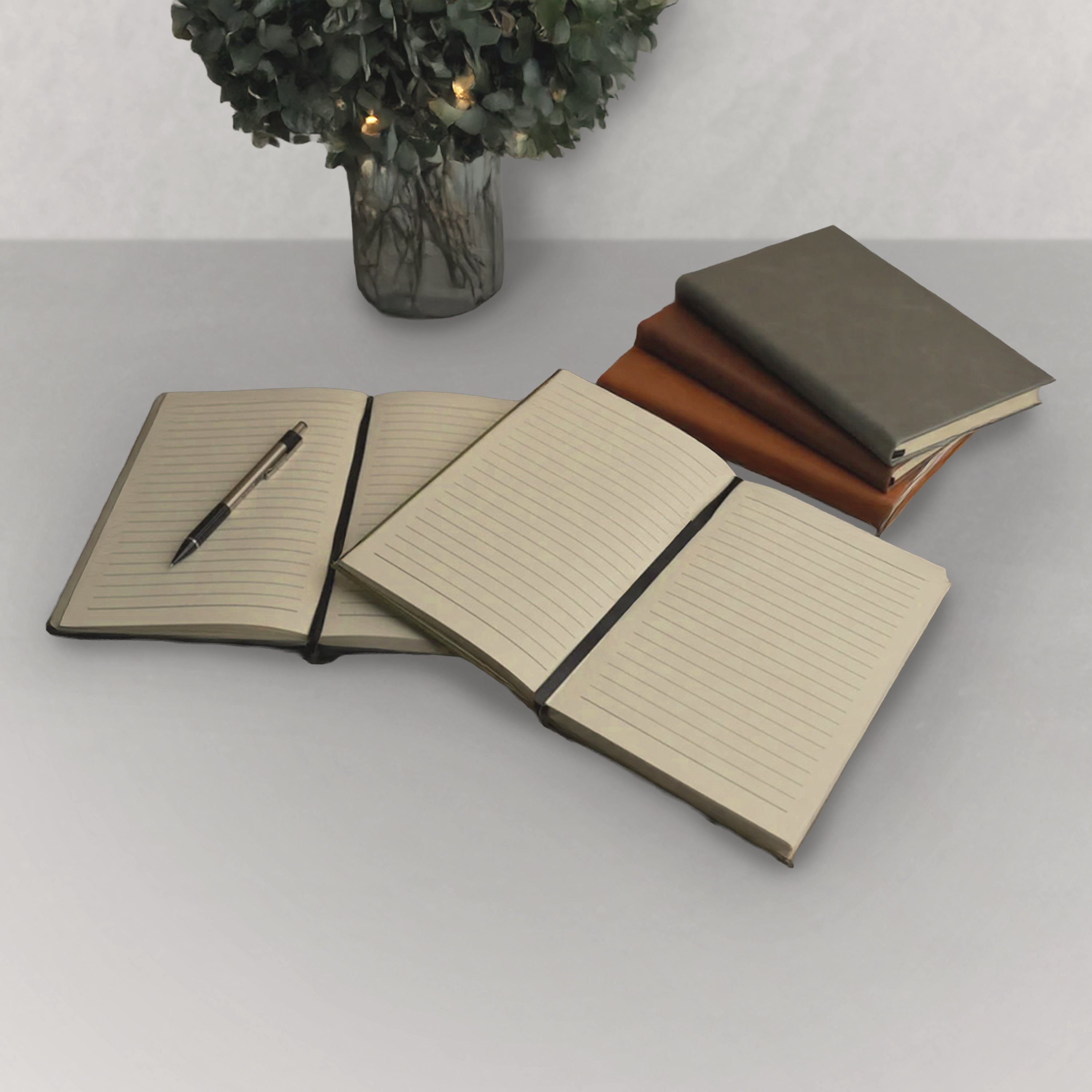 Set of notebooks with a pen on a light gray surface with a vase of flowers in the background.